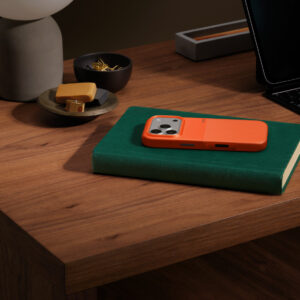 Funda Rugged de Beats, color naranja sierra, acoplada a un iPhone 17 Pro, sobre un libro en un escritorio de madera