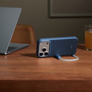 Funda Kickstand de Beats, color azul cobalto, acoplada a un iPhone 17 Pro, parte lateral, sobre una mesa de madera en horizontal, con el conector magnético del cordón acoplado al lateral para sujetar el iPhone